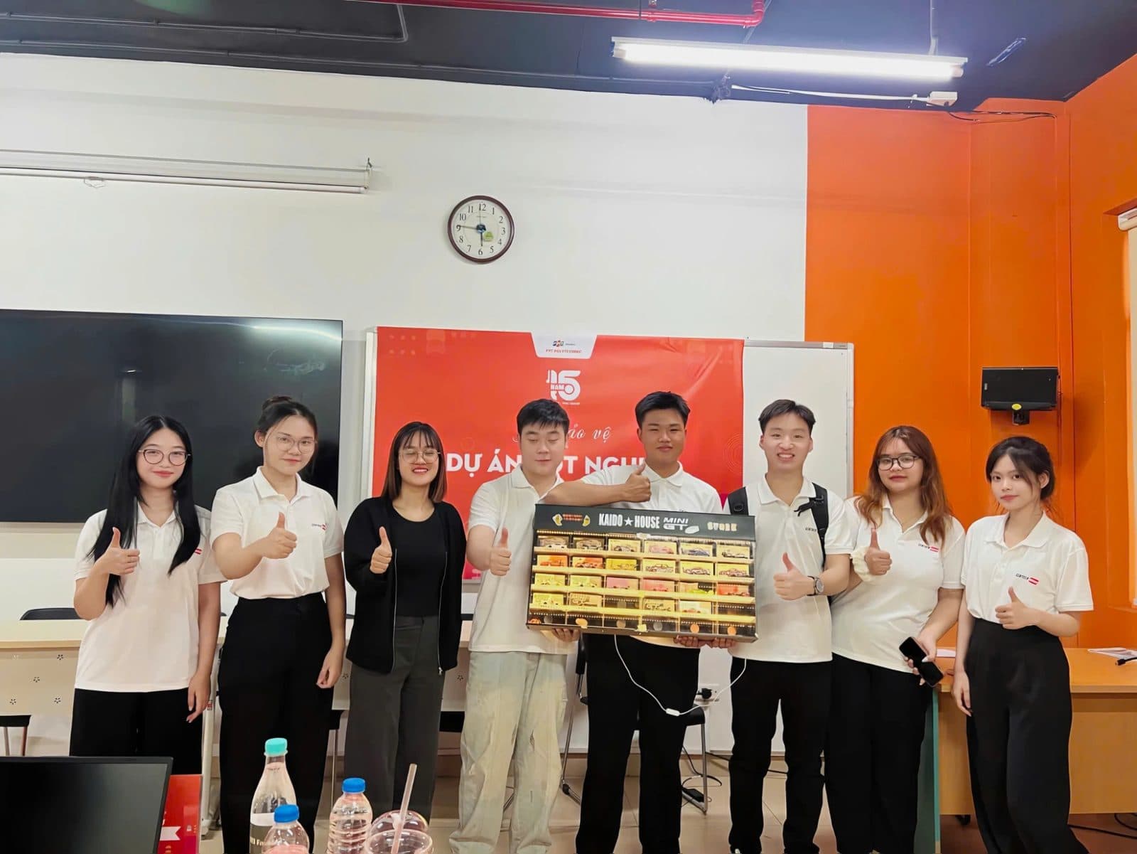 Nhóm sinh viên mặc áo Sentock giơ ngón cái, cầm kệ đèn trưng bày Kaido House × Mini GT tại buổi bảo vệ dự án tốt nghiệp ở FPT Polytechnic